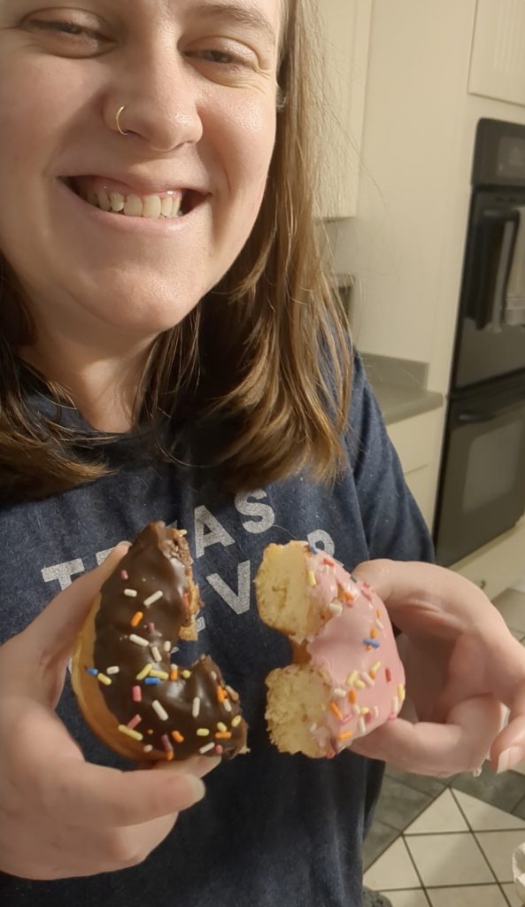 a woman holding half of a chocolate donut with sprinkles and a half of a pink frosting donut with sprinkles. The two halves are held together to resemble a full donut
